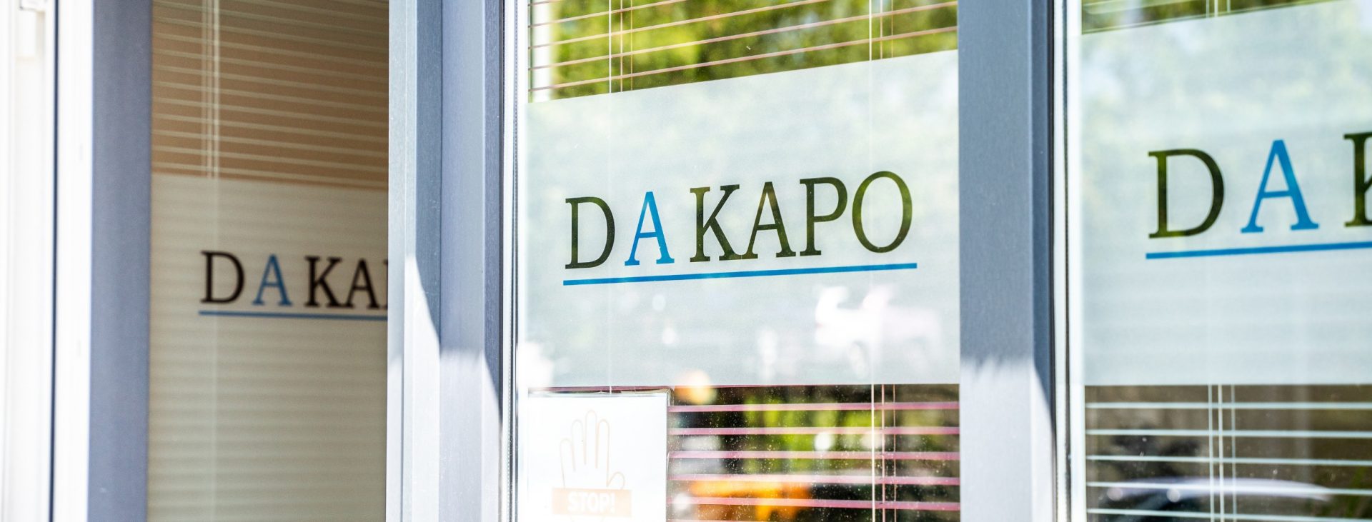 Eingang von DAKAPO mit einer Glastür, die das Straßenbild reflektiert, zeigt das Firmenlogo und ein STOP-Schild für COVID-19-Maßnahmen.